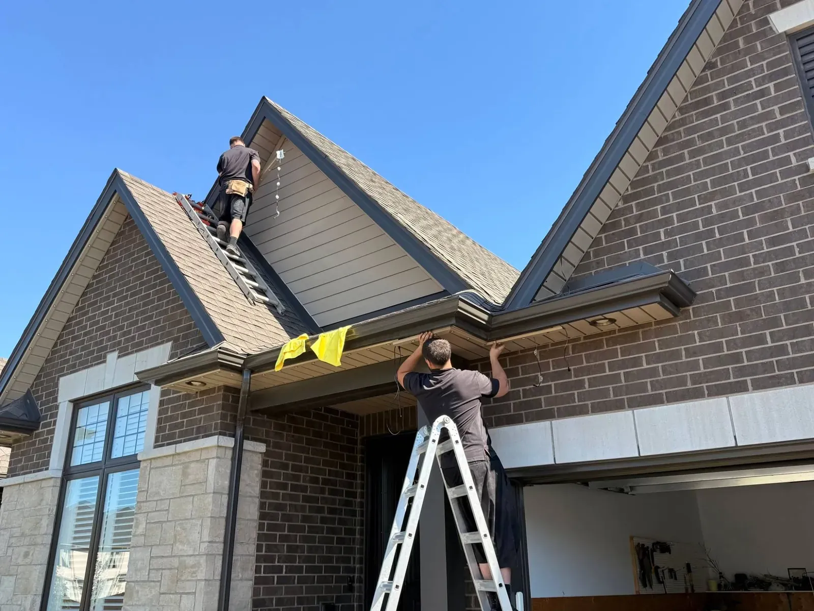 Two installers on ladders mounting permanent lighting track along the soffits and gables of a brick home